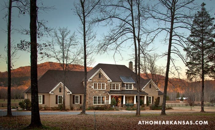 Large stone and brown house with a gable roof, surrounded by trees and hills.
