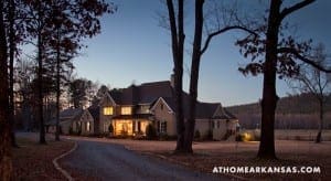 Large stone house at dusk, surrounded by trees and a gravel driveway.