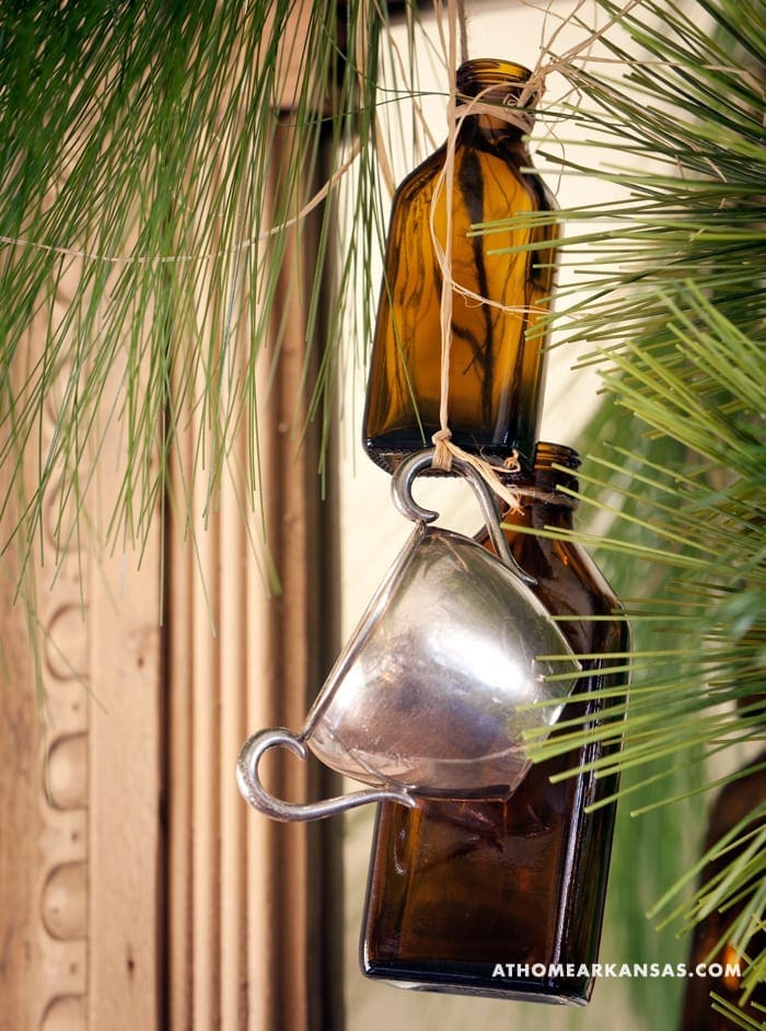 Brown bottle and silver teacup hanging from pine branches.