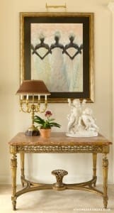 Marble console table with ornate gold accents, artwork, lamp, and sculptures.