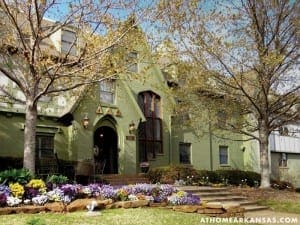 Green Victorian house with flower beds and trees.