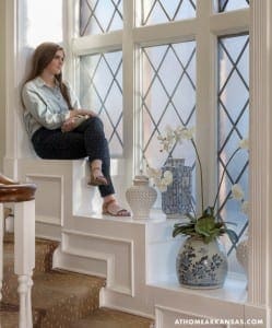 Woman seated on a staircase with a window, orchid, and decorative vase.