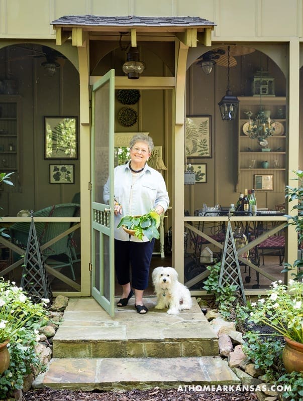 Woman holding plants with a small dog on a porch.