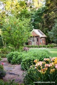 Small, green cottage with a metal roof surrounded by flowers and shrubs.