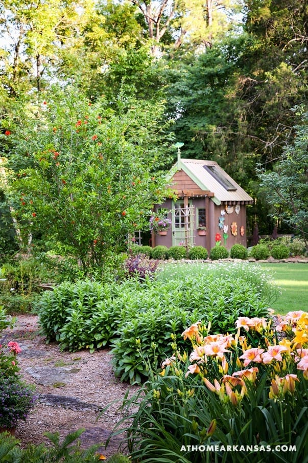 Small, green cottage with a metal roof surrounded by flowers and shrubs.
