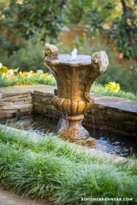 Stone fountain with a sculpted urn, flowing water, and lush greenery.