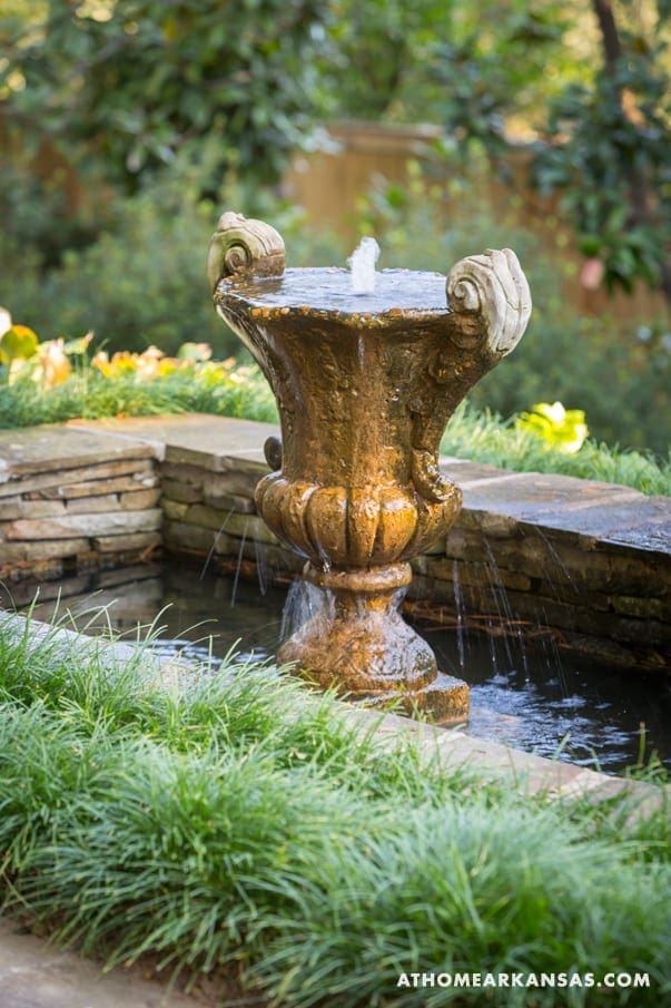 Stone fountain with a sculpted urn, flowing water, and lush greenery.