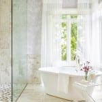 White bathtub in a marble and glass bathroom with sheer curtains.