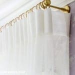 White linen curtains with gold rod on purple wallpaper.