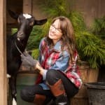 Woman in plaid shirt and boots petting a goat.