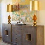 Dark wood console table with two gold lamps and abstract art.