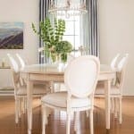 White dining table with upholstered chairs, chandelier, and curtains.