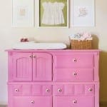 Pink dresser with drawers and floral knobs, featuring framed artwork and a basket.