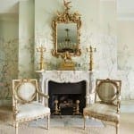 Gold mirror and fireplace with patterned wallpaper and chairs.