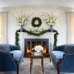 Blue velvet armchairs flank a fireplace with a garland and floral arrangements.