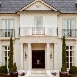 Two-story neoclassical mansion with dark wood entrance, gray shingles, and flanking cypress trees.