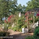 Garden path with flowering bushes, a wooden arbor, birdhouse, and white picket fence.