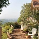 Gravel path leading to a house with a porch overlooking a valley. White chairs sit on the patio.