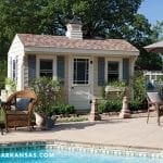 Light yellow shed with blue shutters, cupola, and multi-colored roof. Patio with chairs and pool.