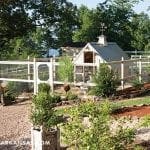 White chicken coop with a weather vane, surrounded by a white fence and lush greenery.