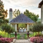 Gazebo with dining table and chairs, stone chimney, and blooming pink flowers.