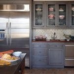Gray kitchen cabinets with glass doors displaying dishware, stainless steel refrigerator, and tiled