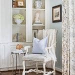White armchair with blue patterned upholstery, light blue pillow, and cream throw. Built-in shelves