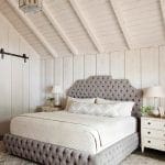 Bedroom with tufted gray bed, white paneled walls, vaulted ceiling, and geometric rug.