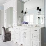 White bathroom vanity with marble countertop, large mirror, and gray upholstered chair.