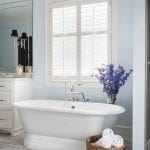 White freestanding bathtub with chrome fixtures, next to a window with white shutters. Blue walls