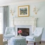 Living room with light blue walls, white fireplace, two light blue armchairs, and patterned ottoman.