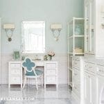 Light blue bathroom with white vanity, ornate mirror, and quatrefoil chair.