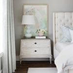 Bedroom with tufted headboard, distressed nightstand, abstract art, and pale blue lamp.