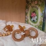Wooden baby toy, pacifiers, and a book titled 'Spring' on a white cloth.