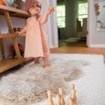 Baby in peach dress and bonnet holding wooden ball near toy bowling pins