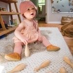 Baby in pink bonnet and dress on fluffy rug with wooden toys.