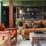 Living room with green walls, wood fluted column, tan sofa, plaid ottoman, and bar area.