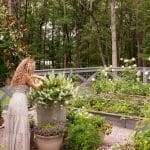 Woman tending to a raised garden bed with white flowers and purple plants.