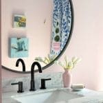 Pink wall with round mirror, floral arrangement, and black faucet.