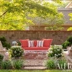 Outdoor seating area with red cushions and wooden chairs, surrounded by greenery and a brick wall.