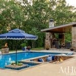 Outdoor pool area with stone fireplace, blue umbrella, and landscaping.