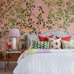 Pink wall with floral wallpaper, bed with patterned pillows and a lamp.