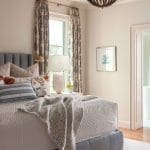 Gray upholstered bed with patterned curtains and a beaded chandelier.