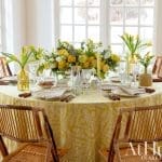 Dining table set with yellow floral tablecloth, yellow tulips, and bamboo chairs.
