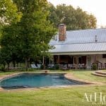 Farmhouse with metal roof, stone chimney, and covered patio overlooking pool and lawn.