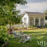 White garden shed with large windows, Adirondack chairs, and picket fence