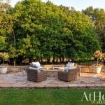 Stone patio with wicker furniture around a fire pit, surrounded by trees and flowers.