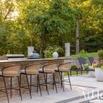 Outdoor kitchen with stone counter, bar stools, grill, and lounge chairs.
