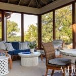 Sunroom with blue sofa, wicker chairs, and white tables, overlooking trees.
