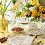 Yellow floral tablecloth with place settings, yellow tulips, and a floral arrangement.